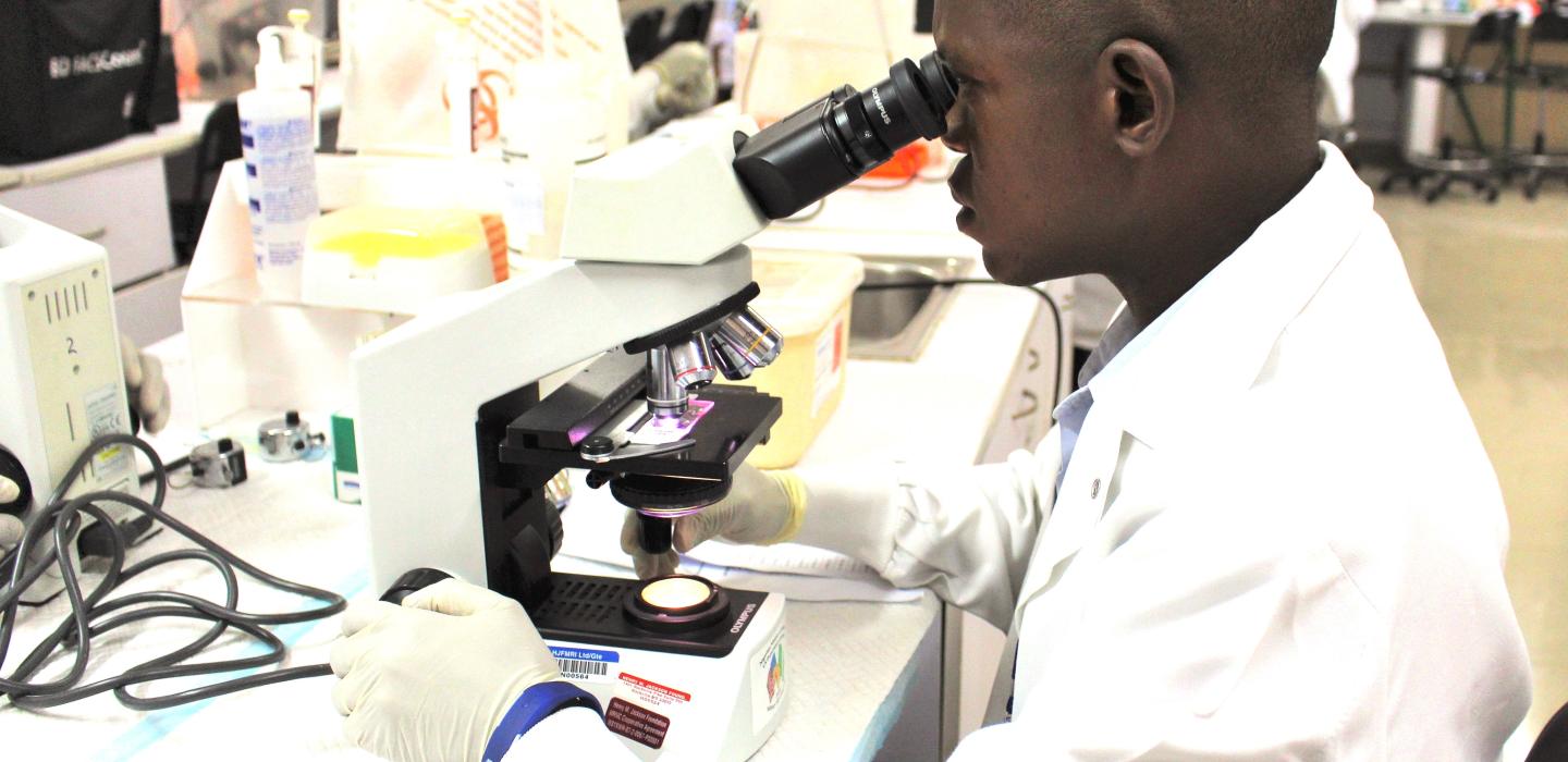 A man in a white lab coat looks into a microscope.