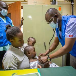 Two masked health workers examine a young girl sitting on her mother's lap.